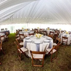 A tented wedding reception set by All About Events, with round tables and wooden folding chairs on grass