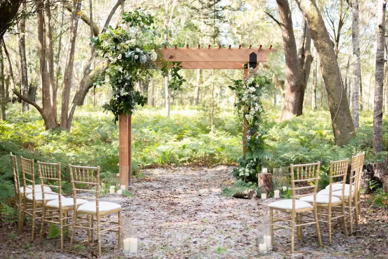 Wooden ceremony arbor dressed in white florals and greenery, with gold chiavari chairs set on a sand clearing surrounded by ferns and palms at Fern Oak Estate