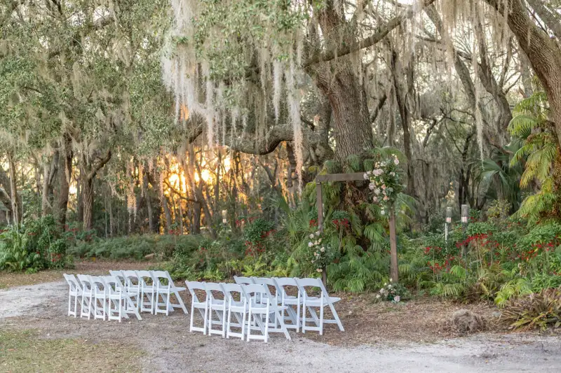 Two blocks of white folding chairs facing a simple wooden arbor with blush florals, under live oaks hung with Spanish moss at sunset at Fern Oak Estate in Pomona Park Florida