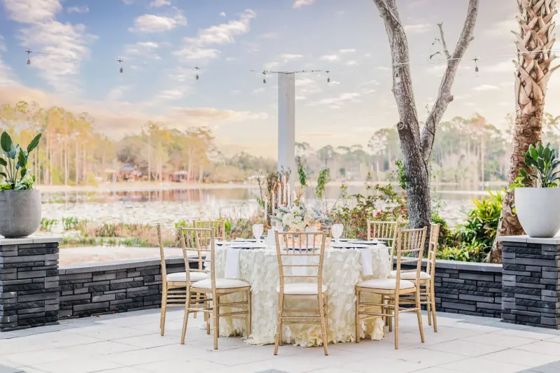 Round table with gold chiavari chairs on the paver terrace at Fern Oak Estate, string lights overhead and the lily-covered lake beyond a low stone wall