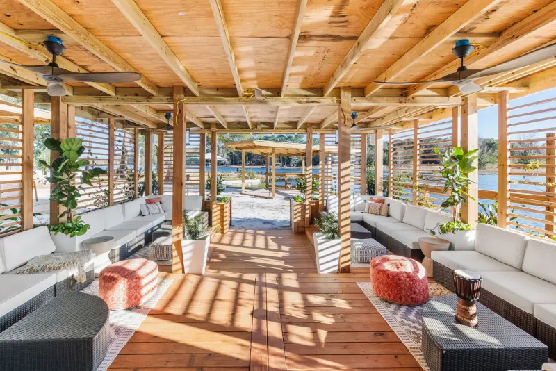 Open-air lakeside pavilion at Fern Oak Estate with a timber ceiling, ceiling fans, slatted wood walls and sectional lounge seating opening onto sand and the lake