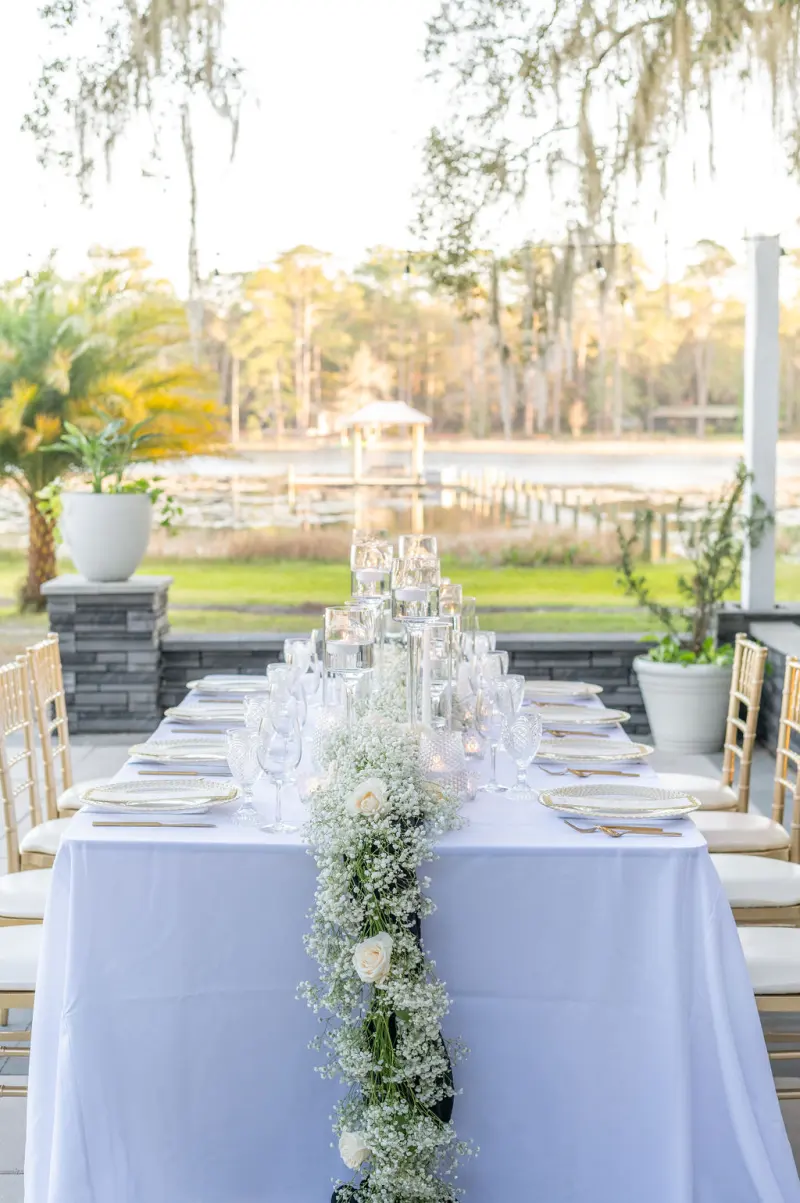 Long reception table with a babys breath runner and gold chiavari chairs on the covered terrace at Fern Oak Estate, looking out to the spring-fed lake and a lakeside gazebo