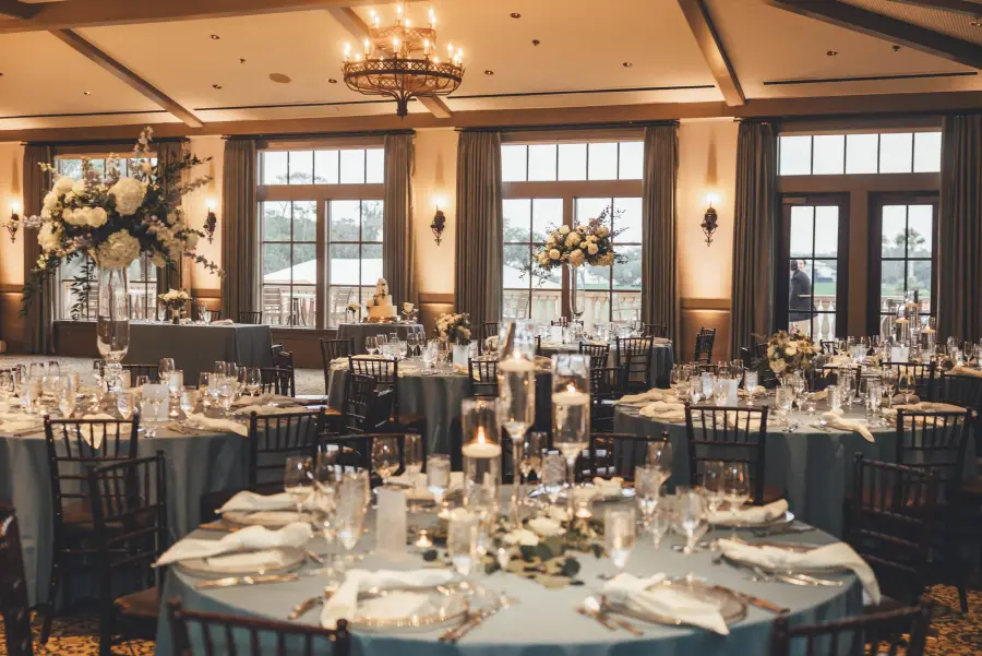 A reception room set by First Coast Weddings and Events, with tall white florals, blue linens, dark chiavari chairs and a chandelier, before guests arrive