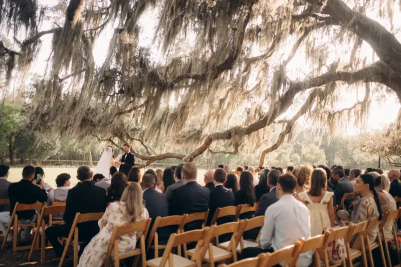 Wedding guests seated in wooden folding chairs beneath a sprawling live oak limb during a pasture ceremony at Golden Creek Ranch