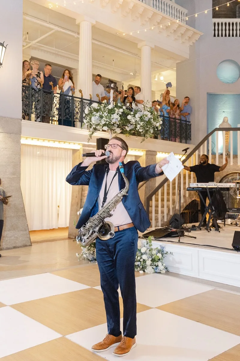 Corey Peterson emceeing a Lightner Museum wedding with a microphone in one hand and a saxophone slung across his shoulder