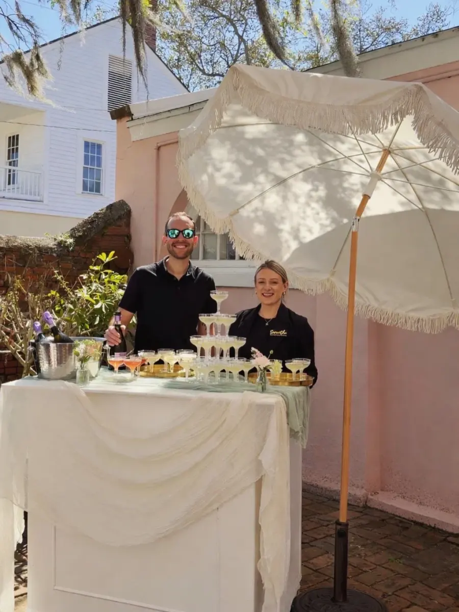 A draped white Maci's Pour bar set up outdoors under a fringed umbrella, with a coupe glass champagne tower and chilled bottles on ice