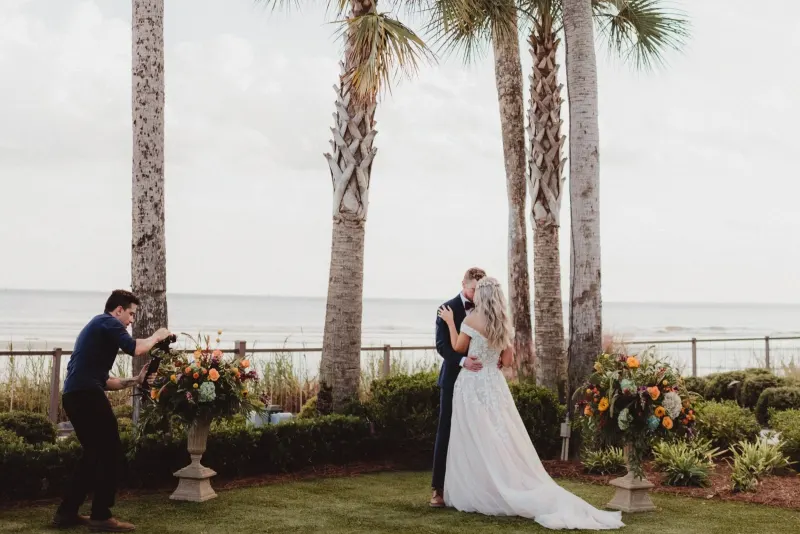 A Rene Visuals cinematographer filming a couple kissing at an oceanfront Florida wedding ceremony framed by palm trees