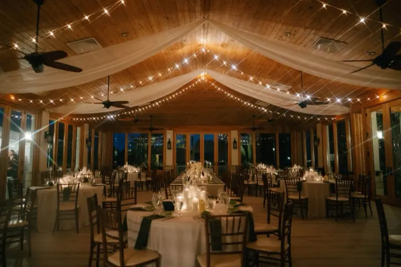 A wedding reception under ceiling draping and strings of warm cafe lights installed by St. Johns Illuminations, with round tables and chiavari chairs in a wood-ceilinged room at night