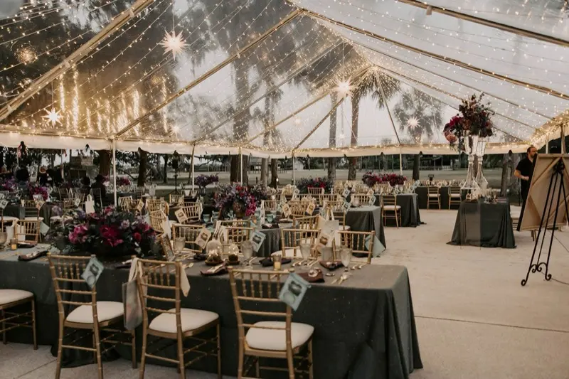 A clear-top tent strung with fairy lights by St. Johns Illuminations, over long tables with dark linens and gold chiavari chairs at dusk