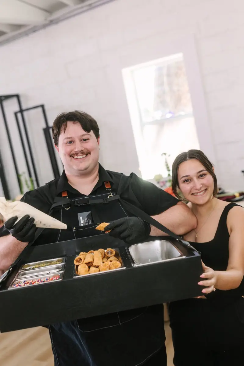 Anthony and Sarah of That Cannoli Guy, Anthony wearing the harness tray of filled cannolis and a black apron