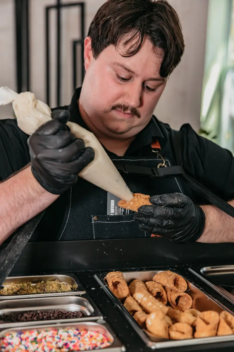 Anthony of That Cannoli Guy piping filling into a cannoli shell over a tray of shells and toppings